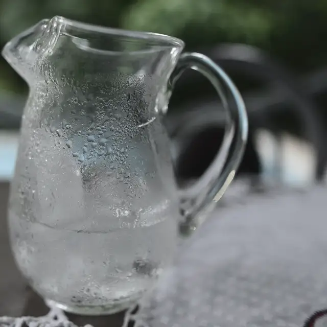 Güçlü yan ışıkta dış yüzeyi terlemiş boş bir cam sürahi, yanında devrilmiş boş bir bardak ve masada dağınık birkaç su lekesi.