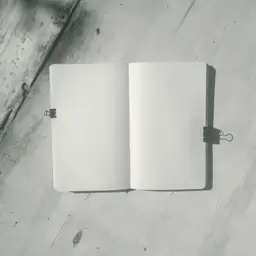 Gri betonun üzerinde açık duran bir defter; iki sayfası da bomboş, dış kenarlarını iki siyah kıskaç tutuyor.