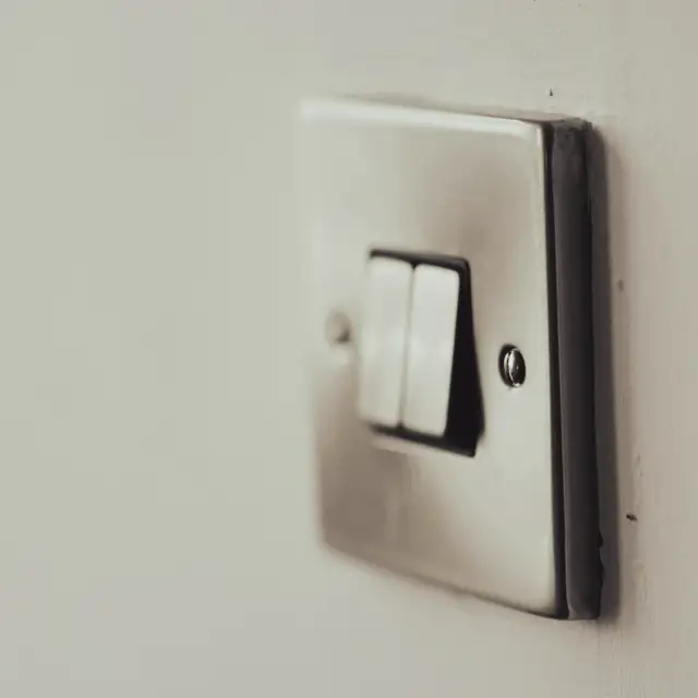 Two white electrical switches set into a single brushed metal plate on a pale wall; there is no writing or number on them.