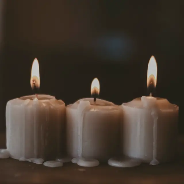 Koyu zeminde yan yana yanan üç kalın mum, üçü de farklı seviyeye kadar erimiş; gövdelerinde donmuş mum akıntıları var.