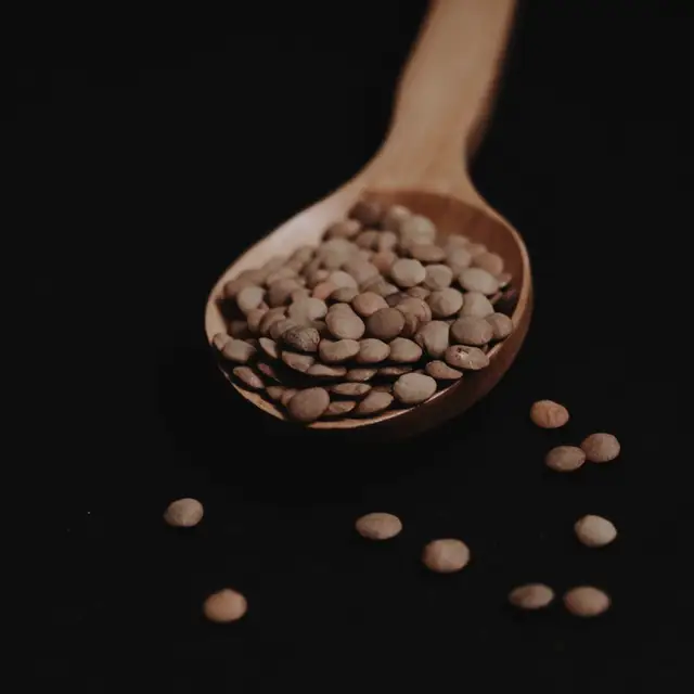 Tamamen siyah zeminde ahşap bir kaşık dolusu mercimek; kaşığın önüne bir avuç tane saçılmış, taneler tek tek sayılabiliyor.