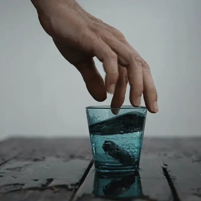 Koyu ahşap bir tezgâhta duran mavi bardağa uzanan bir el; arka plan sığ alan derinliğinde eriyor.