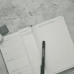An open spiral notebook lies on pale gray concrete, its left page printed as a ruled list and its right page nearly blank with a black pen resting on it.
