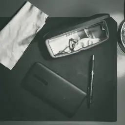 Black and white overhead square of a dark desk: folded eyeglasses in an open case, a phone lying face down below them, a slim pen alongside, a crumpled sheet of paper at the top left, and the rim of a round metal object at the top right.