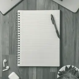 Gri ahşap bir masada, üstünde siyah bir kalem duran açık ve boş çizgili spiral defter; kenarlarda dizüstü bilgisayarın köşesi, kablosuz kulaklıklar ve bir bitki yaprağı görünüyor.